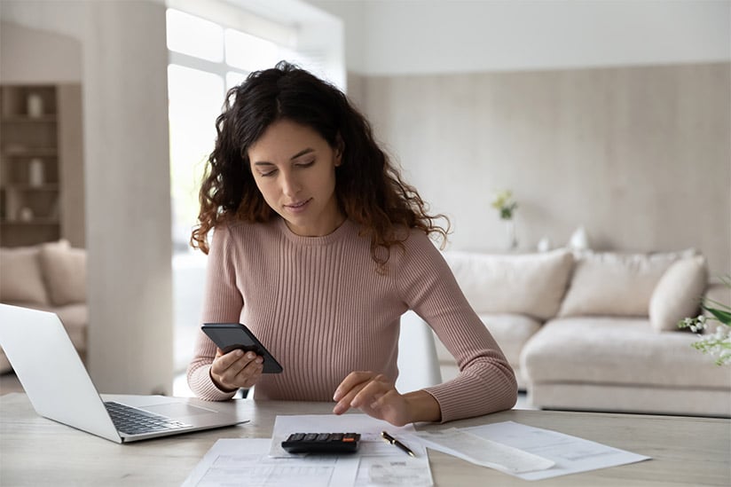 Attractive Latina woman is sitting at a desk at home and managing her household finances and student loans while using a calculator and her smart phone.