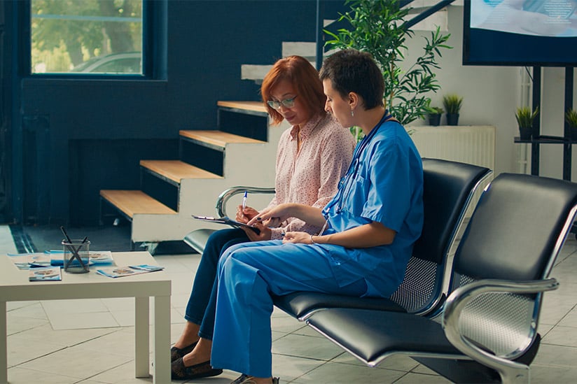 Two nurses, one in scrubs, discuss a patient's chart earnestly, embodying the principle of veracity in nursing through honest communication in a clinic waiting area.