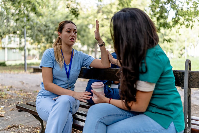 Two nurses in scrubs have a serious conversation on a park bench, one gesturing while holding a coffee cup, the other listens intently. Trees provide a peaceful backdrop.