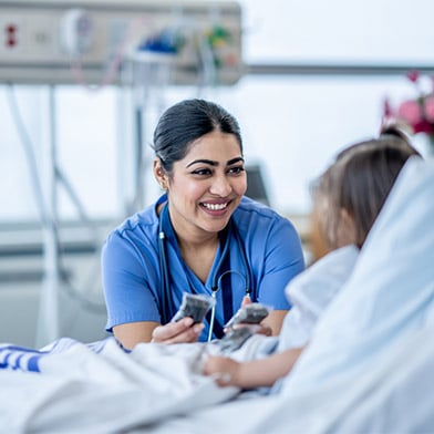 A female nurse leans in closely as she checks on a young patient after surgery. The little girl is wearing a hospital gown and tucked into bed as she talks with her nurse.