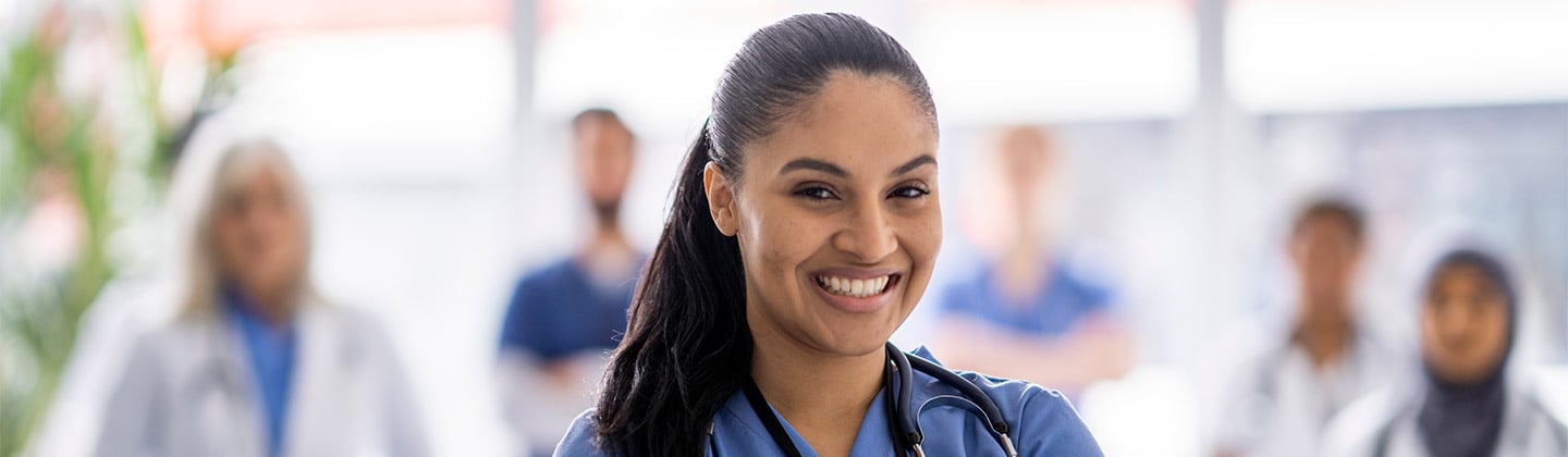 A female bi-racial nurse stands with her arms crossed in front of her and a smile on her face, as she poses for a portrait. She is wearing scrubs, and her team can be seen standing with her in the background.