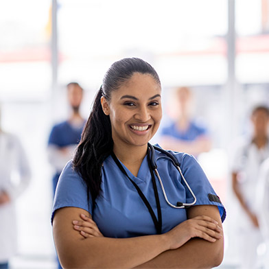 A female bi-racial nurse stands with her arms crossed in front of her and a smile on her face, as she poses for a portrait. She is wearing scrubs, and her team can be seen standing with her in the background.