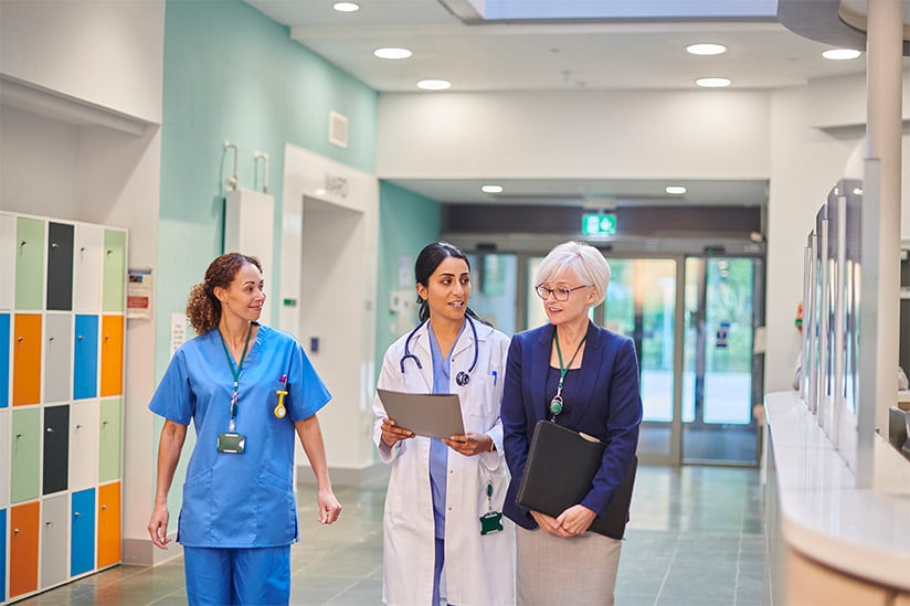 Female utilization nurse walking in a hospital hallway discussing a patient’s care with a female nurse and a female doctor.