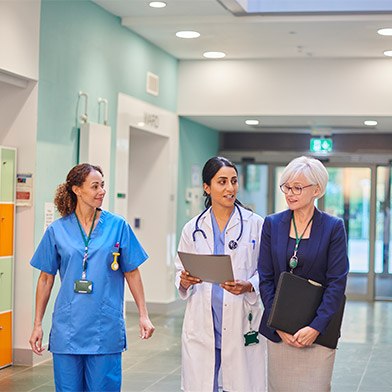 Female utilization nurse walking in a hospital hallway discussing a patient’s care with a female nurse and a female doctor.
