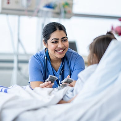 A female nurse of Middle Eastern decent sits at the edge of a hospital bed as she checks in on her young patient. She is wearing blue scrubs and is attempting to cheer the young girl up.