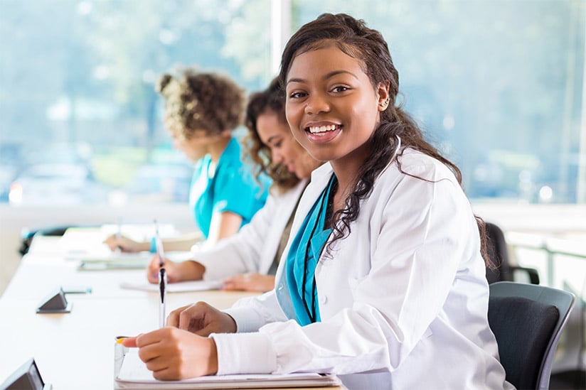 Beautiful African American female nursing student smiles confidently while in class. She is taking notes in a notebook. She is wearing a lab coat over blue scrubs and has dark brown hair.