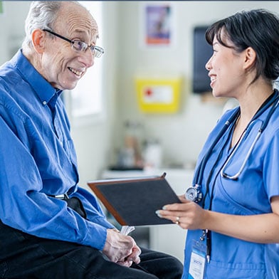 A nurse in a blue scrub top and badge provides coaching to a smiling elderly man in a blue shirt, holding a tablet in a clinic office, demonstrating a patient-centered approach in nursing.