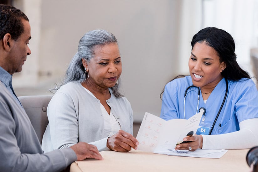 A female African American Adult-Geriatric Nurse Practitioner is seated with an elderly African American woman and going over a treatment plan, while a family member looks on.