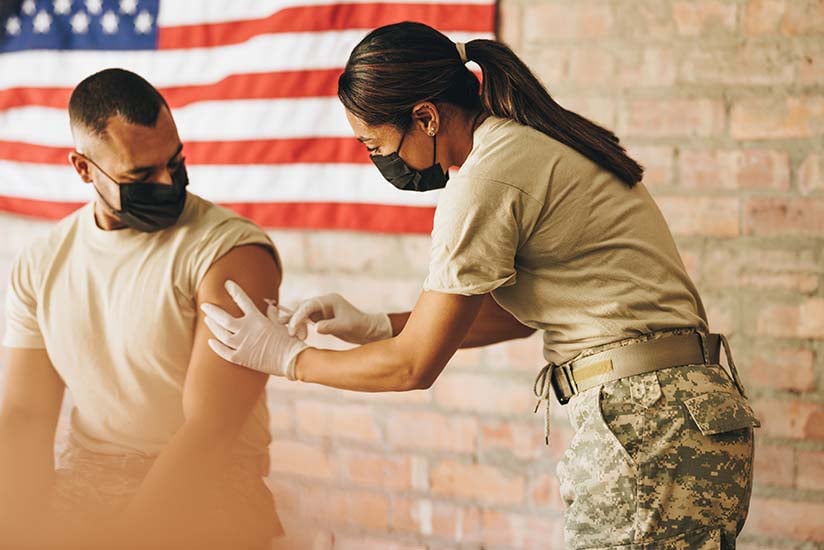 A military nurse in camouflage uniform administers a vaccine to a soldier sitting against an American flag backdrop. Both wear masks, and the focus and precision of the nurse are evident as she performs the injection.