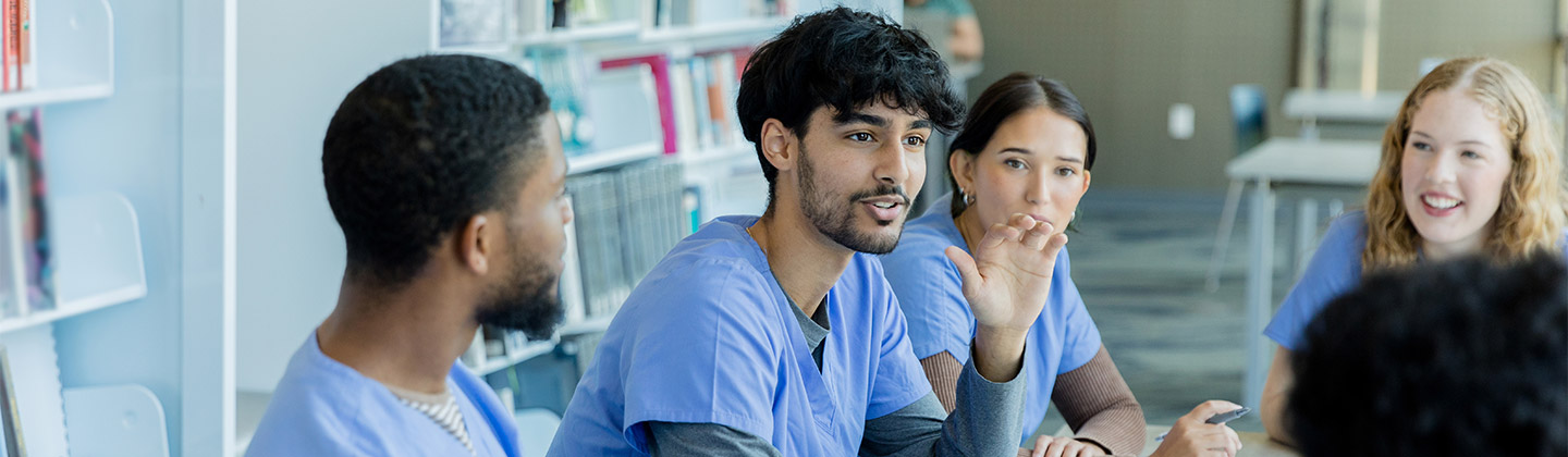 A group of individuals wearing blue scrubs, seated around a table in a bright, modern library setting. The focal point is a man with a beard, gesturing while talking to the group, indicating a discussion or study session in progress. Around him are his colleagues or fellow students, attentive and engaged in the conversation. The environment is casual yet focused, with books and digital tablets visible, suggesting an educational or medical training context.
