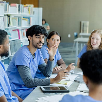 A group of individuals wearing blue scrubs, seated around a table in a bright, modern library setting. The focal point is a man with a beard, gesturing while talking to the group, indicating a discussion or study session in progress. Around him are his colleagues or fellow students, attentive and engaged in the conversation. The environment is casual yet focused, with books and digital tablets visible, suggesting an educational or medical training context.