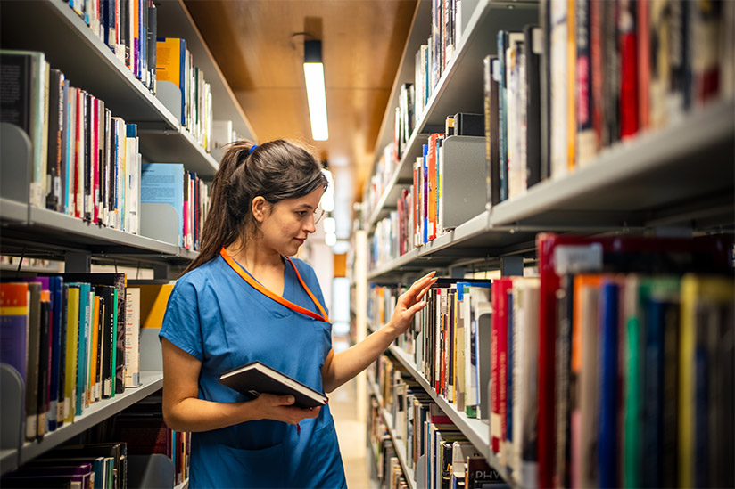A woman in blue scrubs, standing in a library aisle. She is focused on selecting a book from a shelf, holding another book in her hand, indicative of research or study. The rows of colorful book spines suggest a vast collection of resources, and the overall ambiance is quiet and studious. Her attire and the context imply that she is a nurse or a medical student in the midst of academic pursuits.