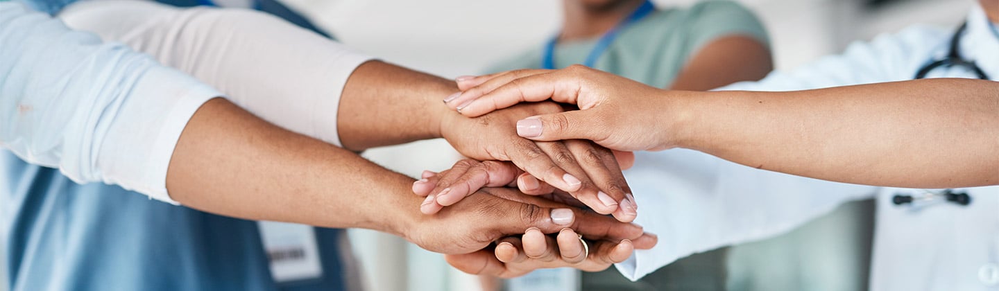 A group of healthcare workers are standing in a circle and each person has an arm outstretched toward the center of the circle. Their hands are stacked on top of each other in a supportive manner indicating they are a team. Their faces are not in focus, but they appear to be smiling.