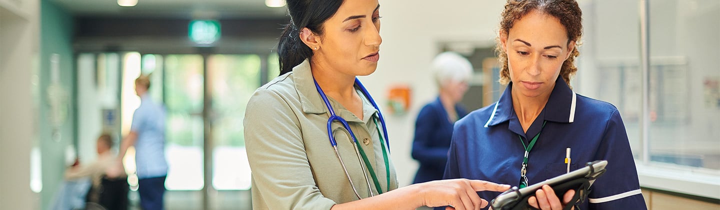 This image portrays two healthcare professionals in a hospital corridor. A woman in a nursing uniform, with a stethoscope around her neck, attentively reviews information on a digital tablet held by a colleague in business attire. Their expressions and body language suggest a serious discussion, indicative of nursing leadership and management in action. The bustling background with other healthcare staff and patients emphasizes the dynamic hospital setting.