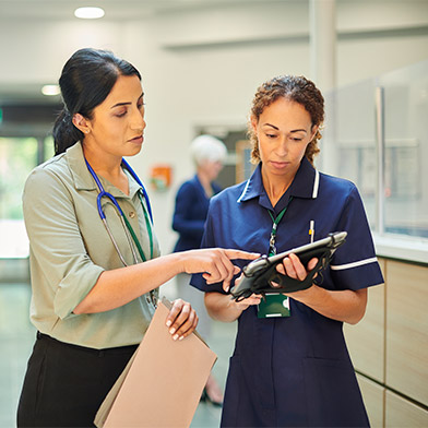 This image portrays two healthcare professionals in a hospital corridor. A woman in a nursing uniform, with a stethoscope around her neck, attentively reviews information on a digital tablet held by a colleague in business attire. Their expressions and body language suggest a serious discussion, indicative of nursing leadership and management in action. The bustling background with other healthcare staff and patients emphasizes the dynamic hospital setting.