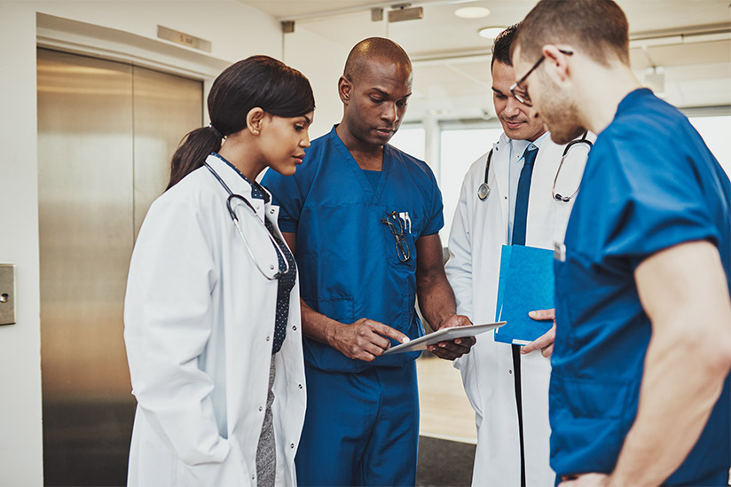 This image shows a diverse group of healthcare professionals, including nurses and doctors, huddled around a clipboard. The focus is on a nurse leader, standing out in blue scrubs, actively engaging with the team. He, along with his colleagues in white coats, appears to be discussing patient care or medical procedures. The group's concentrated demeanor and the clinical environment underscore the collaborative nature of nursing leadership.