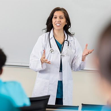 A nurse educator in mid-lecture, standing confidently in front of a classroom. She is dressed in a white lab coat with a stethoscope around her neck, gesturing openly to her students. Her expression is enthusiastic and engaging, suggesting an interactive teaching moment. The blurred foreground shows the back of students’ heads, focusing attention on the educator.