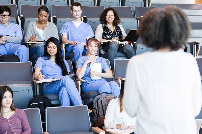 This image presents a nurse educator from the students' perspective, standing at the front of a lecture hall filled with nursing students. The students, wearing blue scrubs, are seated in tiered rows, some writing notes and others looking towards the educator, whose back is to the camera. The room has a collegiate atmosphere, with students appearing attentive and the educator actively engaging with the class.