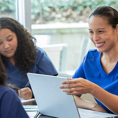 This image captures a cheerful nurse coach in blue scrubs engaging with a student or colleague across a table. She is smiling, gesturing with her hands, illustrating a point or giving advice. A laptop is open in front of her, suggesting a learning or mentorship session. The focus on her face and the attentive gaze of the young woman listening reflects a supportive educational interaction in a bright, welcoming setting.