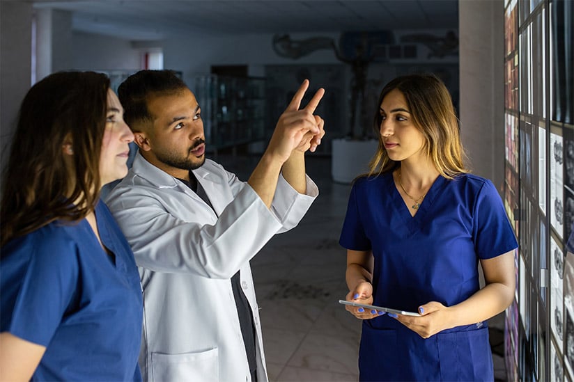 This image features a nurse in blue scrubs holding a tablet, receiving guidance from a healthcare professional in a white lab coat. The coach is pointing upwards, possibly explaining something displayed on a digital screen or board out of the frame. Their expressions are serious and focused, indicative of an in-depth coaching moment. The dim lighting and the clinical background give the impression of an on-site learning scenario within a healthcare facility.