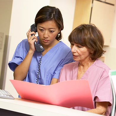 Two nursing professionals wearing scrub outfits are at a desk area in a hospital. One nurse is standing and talking on the phone. The other nurse is seated and holding a folder. Both nurses are looking at information in the folder.