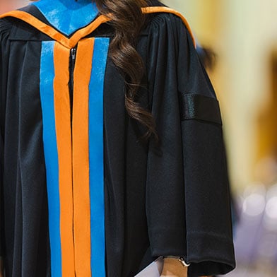  Image of a doctoral student standing while wearing their black graduation gown which has an orange and blue hood and front detail. Only their body is visible. 