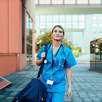 A confident and smiling female nurse wearing blue scrubs is walking outside a hospital on her way to work. She is carrying a blue duffle bag with all the things she will need for a long shift at work.  