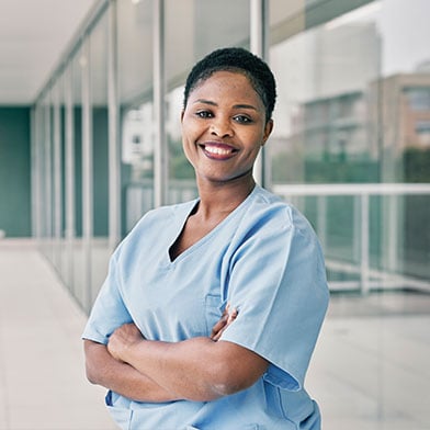 A smiling female nurse is standing in front of her hospital with her arms crossed. 