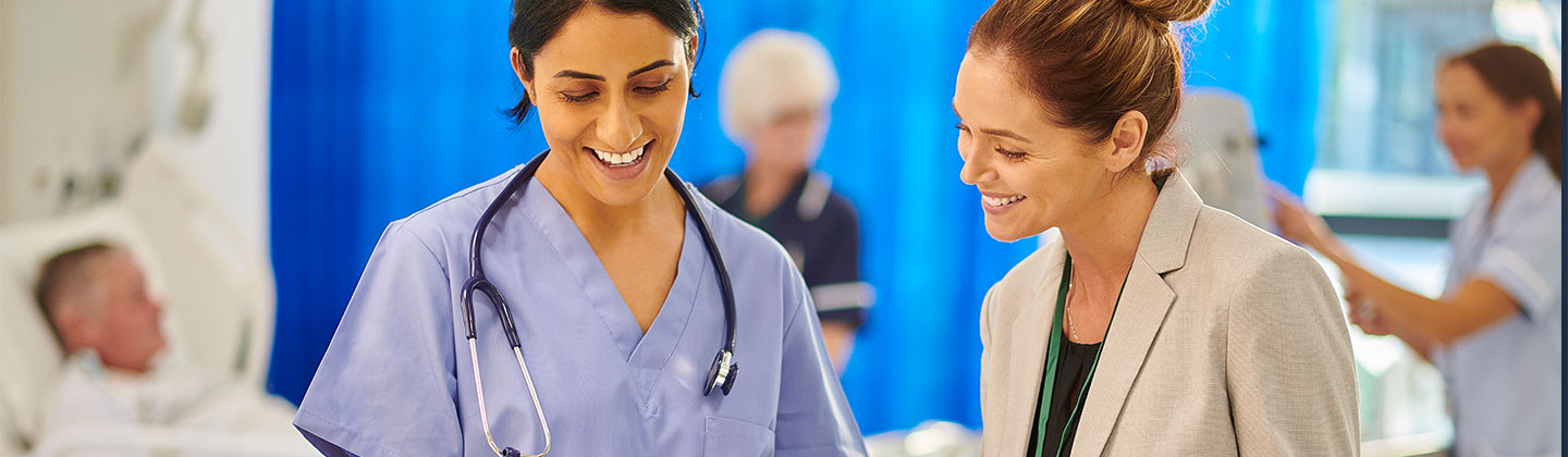A confident female nurse manager is in a patient care area, and she is meeting with a staff member. Both are smiling and looking at some documentation the staff member is holding, and they are engaged in conversation. 