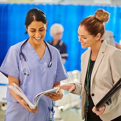 A confident female nurse manager is in a patient care area, and she is meeting with a staff member. Both are smiling and looking at some documentation the staff member is holding, and they are engaged in conversation.  